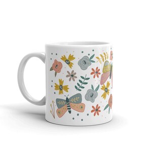 May include: White ceramic mug decorated with colorful butterflies and floral patterns. The design includes butterflies in shades of pink, yellow, and blue, along with various flowers and green foliage. The mug has a white handle and a glossy finish.