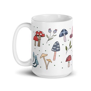 May include: White ceramic mug featuring a whimsical mushroom design. The design includes various colorful mushrooms, green leaves, and small blue berries. The mug has a large handle and is suitable for hot or cold beverages.