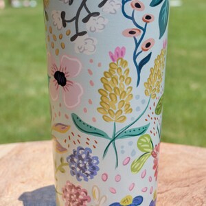 May include: A floral-patterned tumbler with a light blue background. The design features pink, yellow, and blue flowers with green leaves. The tumbler has a dark blue lid and is sitting on a wooden surface.