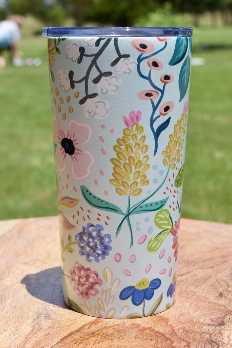 May include: A floral-patterned tumbler with a light blue background. The design features pink, yellow, and blue flowers with green leaves. The tumbler has a dark blue lid and is sitting on a wooden surface.