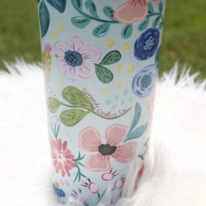 May include: A floral-patterned tumbler with a light blue background. The tumbler is adorned with pink, blue, and white flowers, green leaves, and the text "Simonds Creative Co." The tumbler has a blue lid and is set on a white, fluffy surface.