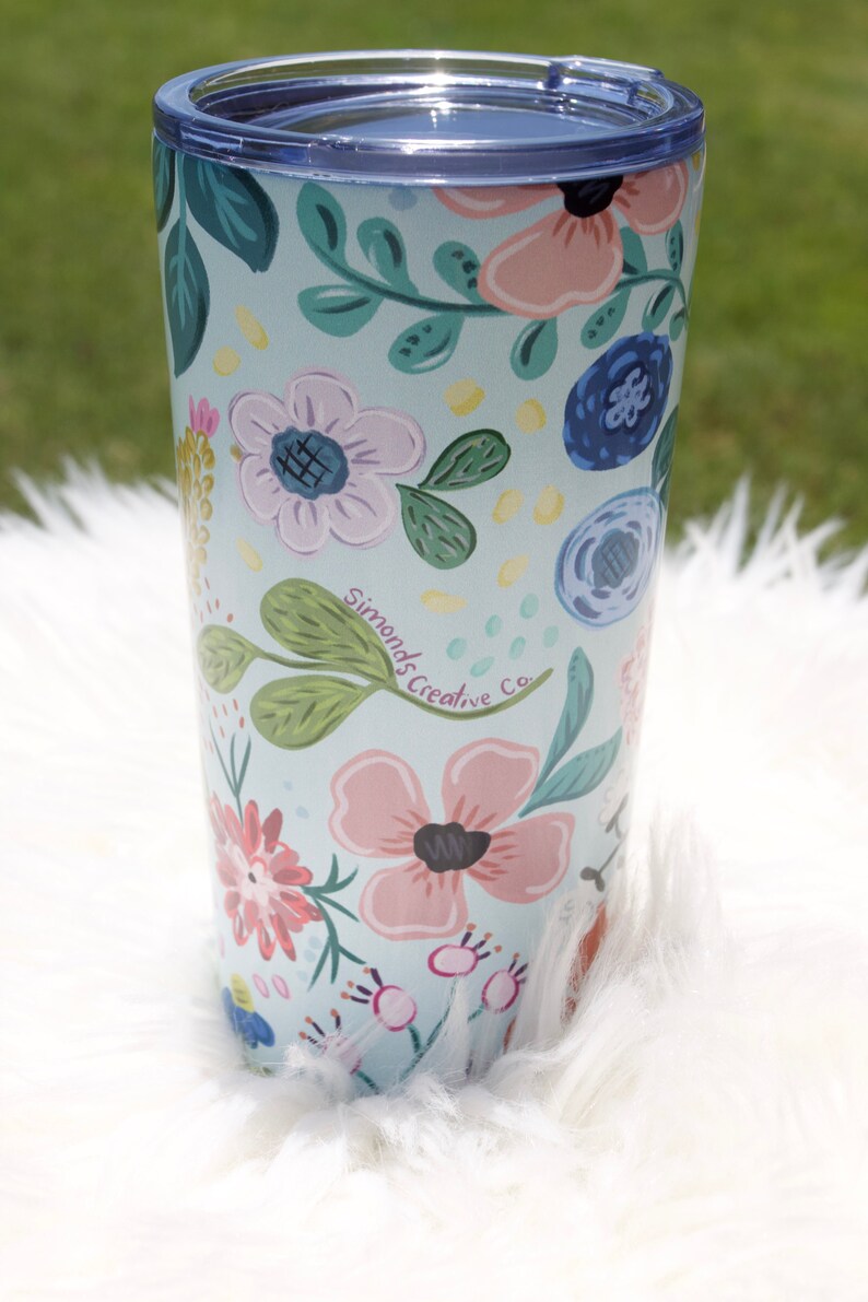 May include: A floral-patterned tumbler with a light blue background. The tumbler is adorned with pink, blue, and white flowers, green leaves, and the text "Simonds Creative Co." The tumbler has a blue lid and is set on a white, fluffy surface.