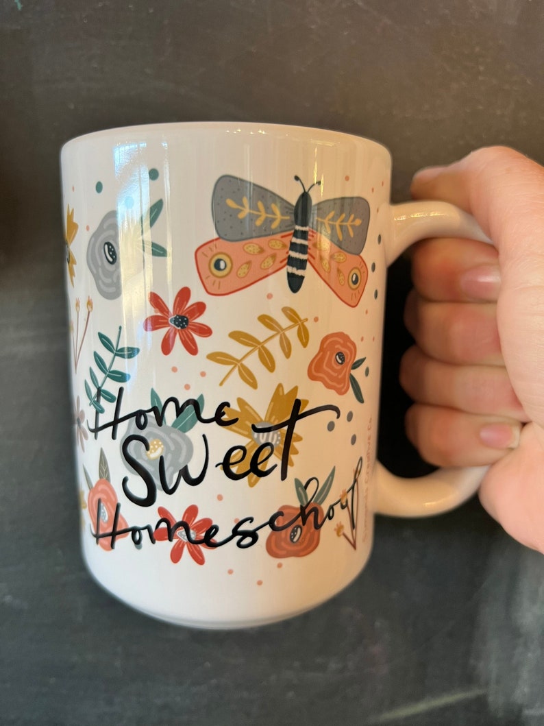 May include: White ceramic mug with a wraparound floral design and a large butterfly illustration. The mug features the words "Home Sweet Homeschool" in black script. The mug is held by a hand.
