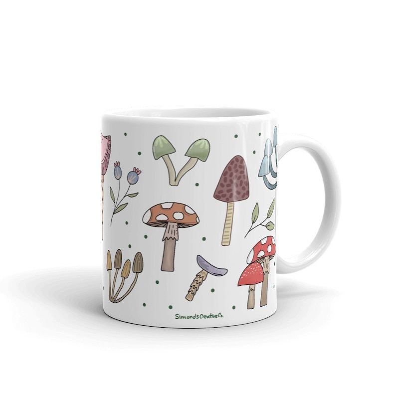 May include: White ceramic mug decorated with colorful illustrations of various mushrooms and foliage. The mug features a handle and is adorned with green polka dots. The text "Simonds Creative Co." is printed on the mug.