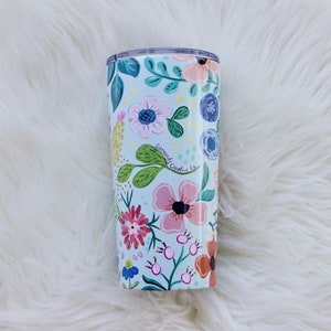 May include: A floral-patterned tumbler with a light blue background. The design features pink, red, and blue flowers with green leaves. The text "Simonds Creative Co." is visible on the tumbler. The tumbler is cylindrical and appears to be made of metal.