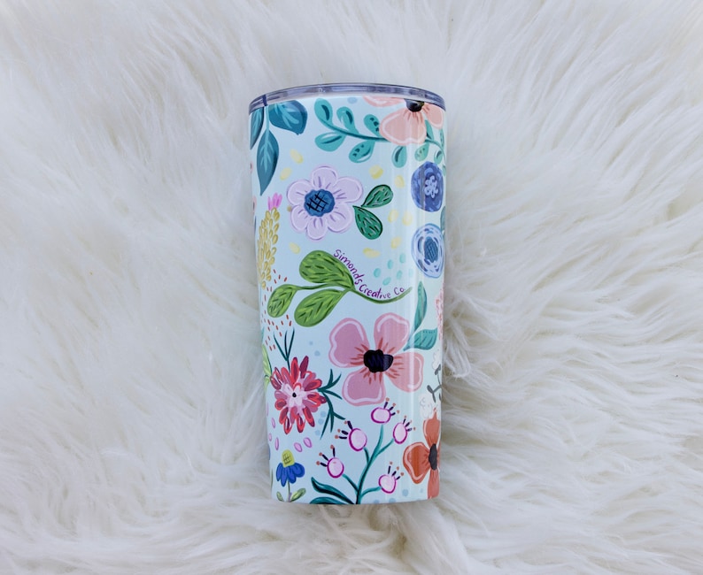 May include: A floral-patterned tumbler with a light blue background. The design features pink, red, and blue flowers with green leaves. The text "Simonds Creative Co." is visible on the tumbler. The tumbler is cylindrical and appears to be made of metal.