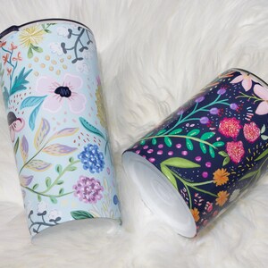 May include: Two floral-patterned tumblers. One is light blue with pink, yellow, and purple flowers. The other is dark blue with pink, orange, and purple flowers. Both have black lids and white interiors. The tumblers are cylindrical.