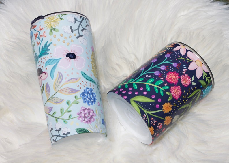 May include: Two floral-patterned tumblers. One is light blue with pink, yellow, and purple flowers. The other is dark blue with pink, orange, and purple flowers. Both have black lids and white interiors. The tumblers are cylindrical.