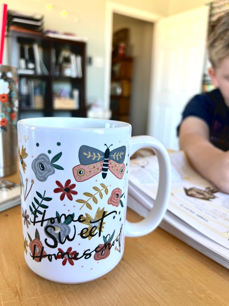 May include: White ceramic mug with a wraparound floral and butterfly design. The mug features a butterfly with pink and blue wings, along with flowers in shades of red, gray, and gold. The words "Home Sweet Homeschool" are written on the mug.