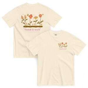 Puede incluir: Dos camisetas de color crema con un diseño floral y el texto "Tend it well" impreso en ellas. La parte delantera de la camiseta tiene un diseño floral más pequeño y la parte trasera de la camiseta tiene un diseño floral más grande.