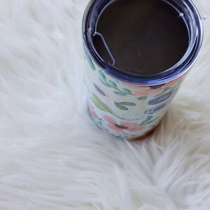 May include: A floral-patterned tumbler with a clear lid. The tumbler features a light blue background with pink, green, and blue floral designs. The lid is clear with a sliding closure. The tumbler is cylindrical and sits on a white, fluffy surface.