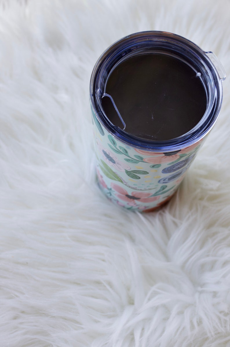 May include: A floral-patterned tumbler with a clear lid. The tumbler features a light blue background with pink, green, and blue floral designs. The lid is clear with a sliding closure. The tumbler is cylindrical and sits on a white, fluffy surface.