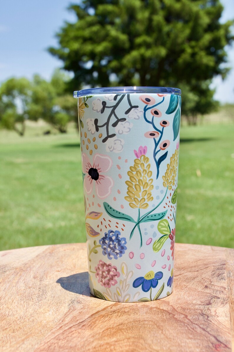 May include: A floral-patterned tumbler with a light blue background. The design features colorful flowers in pink, yellow, and blue. The tumbler has a clear lid and is set on a wooden surface.