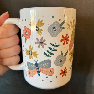 May include: White ceramic mug decorated with colorful floral and butterfly illustrations. The design includes flowers in shades of yellow, peach, gray, and red, along with a butterfly. The mug has a curved handle and is suitable for hot or cold beverages.