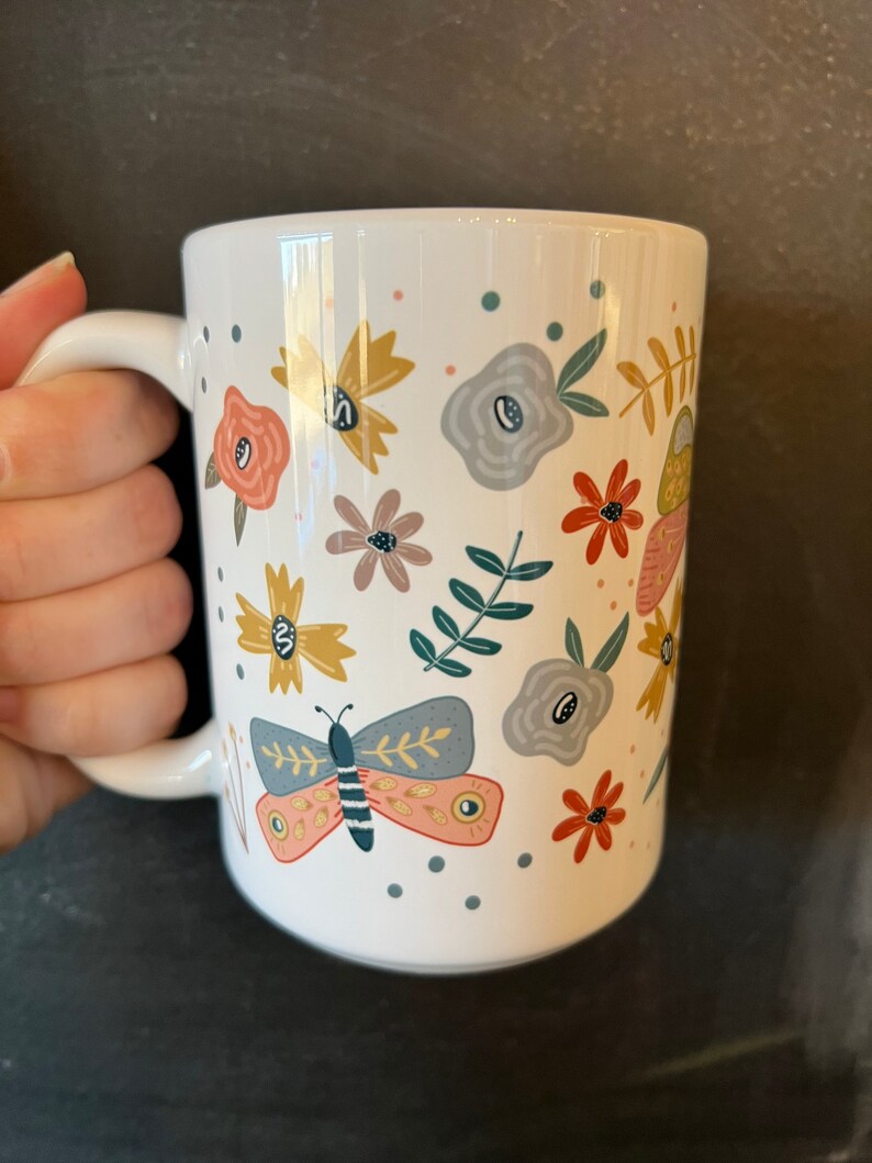 May include: White ceramic mug decorated with colorful floral and butterfly illustrations. The design includes flowers in shades of yellow, peach, gray, and red, along with a butterfly. The mug has a curved handle and is suitable for hot or cold beverages.