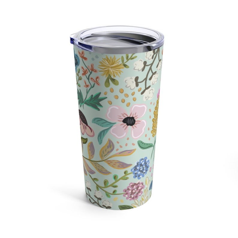 May include: A floral-patterned tumbler with a stainless steel lid. The tumbler features a light green background with a repeating pattern of pink, yellow, blue, and white flowers and green leaves. The tumbler is approximately 7 inches tall.