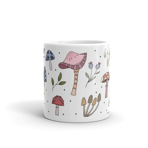 May include: White ceramic mug decorated with colorful mushroom illustrations. The design features various mushroom types in shades of pink, red, brown, and blue, along with green leaves and blue berries. The mug is a standard size for hot beverages.