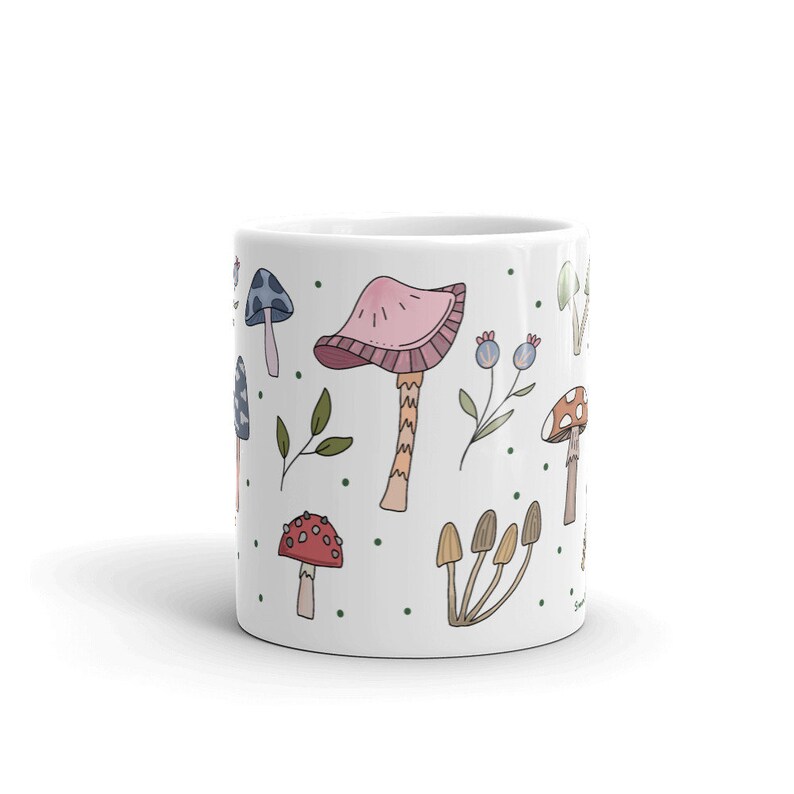May include: White ceramic mug decorated with colorful mushroom illustrations. The design features various mushroom types in shades of pink, red, brown, and blue, along with green leaves and blue berries. The mug is a standard size for hot beverages.