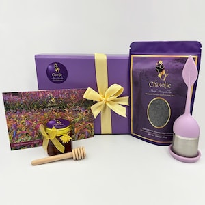 May include: A gift set featuring purple tea, honey, and a tea infuser. The set includes a purple box tied with a yellow ribbon, a jar of honey with a wooden honey dipper, and a bag of tea. A card with a floral image is also included.
