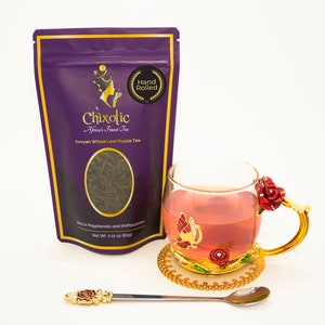 May include: Purple tea leaves in a resealable pouch with the brand name "Chixotic Africa's Finest Tea" and the text "Kenyan Whole Leaf Purple Tea". The pouch is on a white surface next to a glass teacup with a gold rim and a red rose design. The teacup is filled with pink tea and has a gold handle.