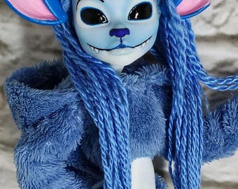 May include: A blue doll with large ears, blue hair, and a blue furry outfit. The doll has a light blue face with black eyes, a purple nose, and a small, smiling mouth. The ears are pink and blue. The doll's outfit is a matching blue color.