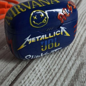 May include: A blue JBL wireless headphone case with a graphic design featuring the band names Nirvana, Metallica, and a smiley face with an 'X' for eyes. The case also has the text 'A.S.S.' and 'Click-It'