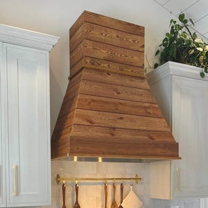 May include: A rustic, wooden range hood with a natural finish. The hood is constructed from horizontal planks of wood, creating a textured, layered appearance. It is installed above a stainless steel range and is flanked by white cabinets with gold hardware.