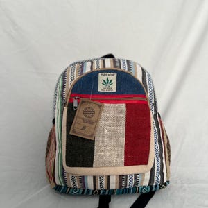 May include: A multi-coloured hemp backpack with a front pocket featuring vertical stripes in black, beige, and red. The top flap is blue with a red zip. The backpack has a tag that says "Handmade in Nepal".