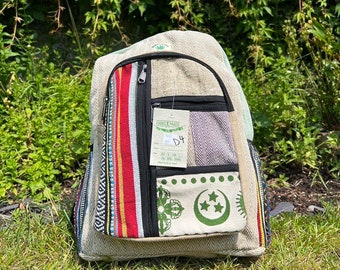 Könnte beinhalten: Beigefarbener Rucksack mit mehreren Taschen und einem Hauptfach mit Reißverschluss. Die Vorderseite hat ein vertikales Streifendesign in Rot, Gelb und Weiß sowie einen Aufnäher mit grünen Symbolen. Die Tasche ist aus gewebtem Material und trägt ein Etikett "HAND MADE".