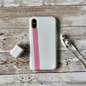 May include: White phone case with a pink stripe and the letter "F" on the bottom right corner. The phone case is on a white wooden surface with white earbuds and a white phone charger.
