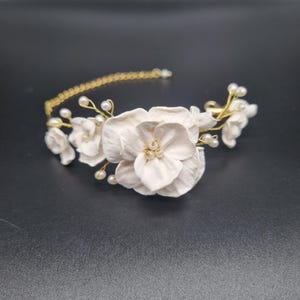 Könnte beinhalten: Ein zartes Blumen-Haarband mit weißen Porzellanblüten und kleinen Perlenakzenten. Die Blüten sind auf einem goldfarbenen Drahtrahmen angeordnet und schaffen ein elegantes und romantisches Accessoire. Ideal für Hochzeiten oder besondere Anlässe.