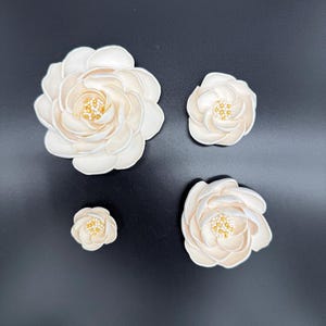 May include: Four ivory-coloured, artificial camellia flowers of varying sizes are arranged on a black surface. Each flower has multiple petals and a centre with small, gold-coloured details. The flowers range from small buds to fully bloomed.