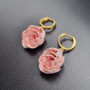 May include: A pair of gold-coloured hoop earrings, each featuring a light pink rose. The roses have a layered petal design, with a darker pink centre. The earrings are set against a dark grey background.