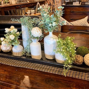 May include: A wooden tray with a variety of white ceramic vases filled with green and white floral arrangements. The tray is sitting on a wooden table.