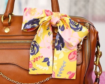 May include: A brown leather handbag with gold-tone hardware, featuring a decorative floral scarf tied to the handle. The scarf has a yellow background with pink and blue floral patterns. A gold chain hangs from the bag.