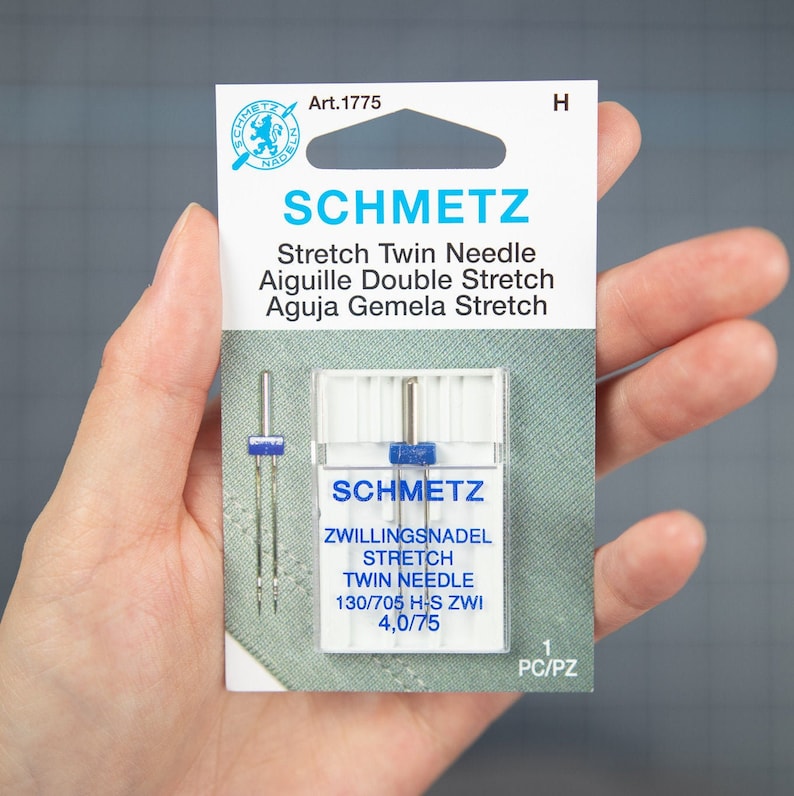 Peut inclure: Un paquet d'aiguilles jumel&eacute;es extensibles Schmetz, taille 130/705 H-S ZWI 4,0/75. L'emballage est blanc avec du texte bleu et un logo bleu et blanc. Les aiguilles sont dans un &eacute;tui en plastique transparent.