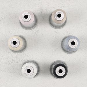 May include: Overhead shot of six spools of thread in various colors: light pink, beige, cream, light blue, white, and black. Each spool is on a clear plastic base, arranged in a circular pattern on a light gray surface.