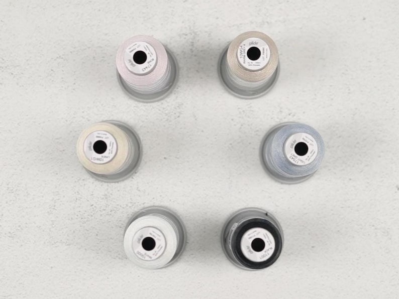 May include: Overhead shot of six spools of thread in various colors: light pink, beige, cream, light blue, white, and black. Each spool is on a clear plastic base, arranged in a circular pattern on a light gray surface.