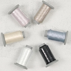 May include: An overhead shot of six spools of thread in various colors: light pink, beige, cream, light blue, black, and white. Each spool is cylindrical with a gray base and top. The threads appear to be shiny and are arranged in a circular pattern.