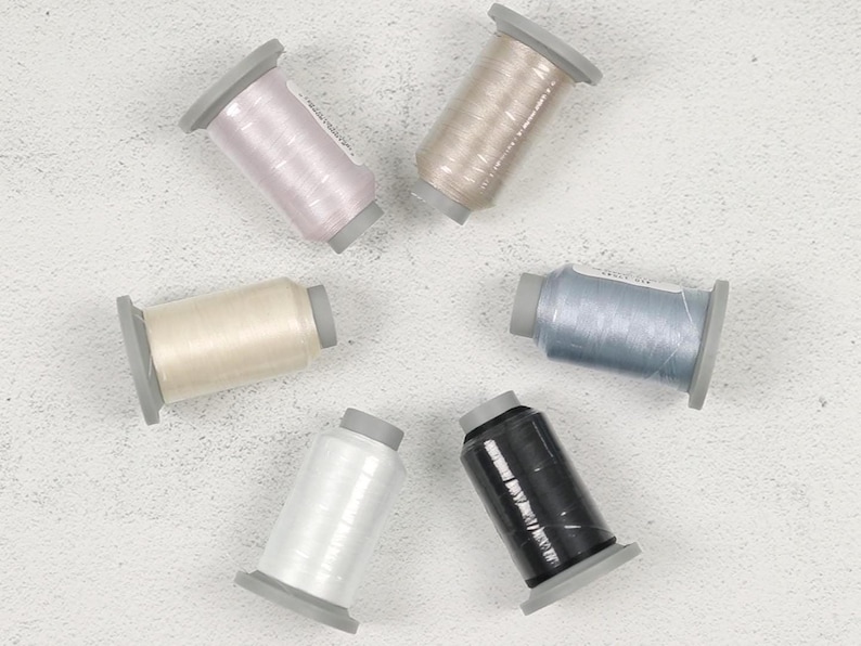 May include: An overhead shot of six spools of thread in various colors: light pink, beige, cream, light blue, black, and white. Each spool is cylindrical with a gray base and top. The threads appear to be shiny and are arranged in a circular pattern.