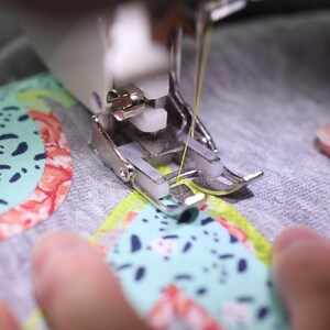 May include: Close-up of a sewing machine needle and foot stitching a colorful fabric with a green and blue pattern. The fabric is being held in place by a hand.