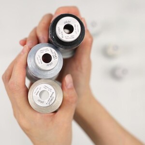 May include: Close-up of three spools of thread in black, light grey, and warm grey. The spools are labeled with the brand "glide" and color information. The thread is tightly wound on the spools, and the image is well-lit.