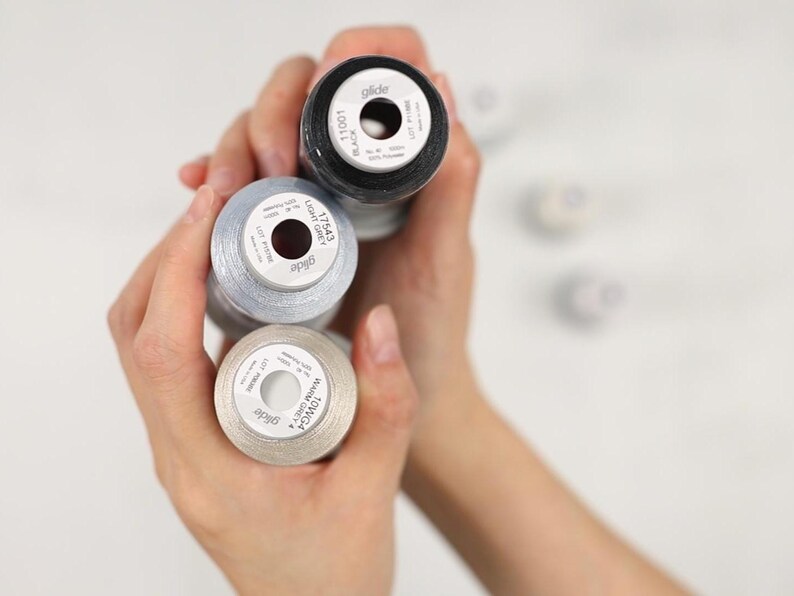 May include: Close-up of three spools of thread in black, light grey, and warm grey. The spools are labeled with the brand "glide" and color information. The thread is tightly wound on the spools, and the image is well-lit.