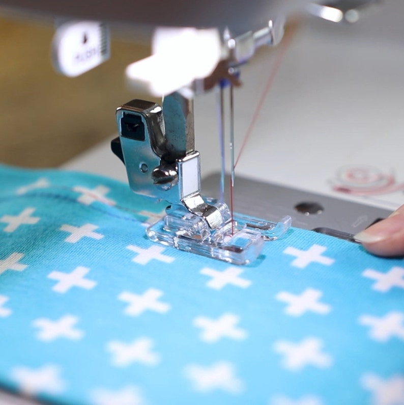 Peut inclure: Gros plan sur l'aiguille et le pied d'une machine &agrave; coudre, cousant un tissu bleu avec un motif de croix blanches.