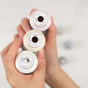 May include: Three spools of Glide brand thread in shades of white and cream are held in a person's hands. The spools have black centers and white labels with text. The thread is likely used for sewing or embroidery projects.