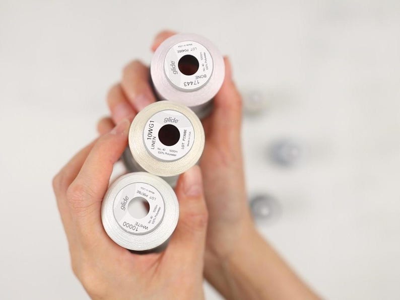 May include: Three spools of Glide brand thread in shades of white and cream are held in a person's hands. The spools have black centers and white labels with text. The thread is likely used for sewing or embroidery projects.