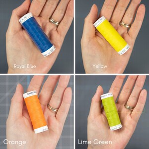 May include: Four spools of thread in different colors: royal blue, yellow, orange, and lime green. The spools are held in a hand.