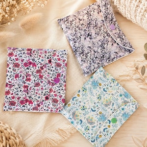 Pode incluir: Três bolsas de tecido quadradas com abas tipo envelope presas por botões de pressão de plástico. Uma bolsa apresenta um padrão floral rosa e vermelho, outra tem uma estampa floral abstrata roxa e cinza, e a terceira mostra um design botânico azul e verde.