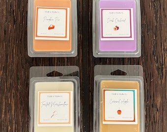 May include: Four rectangular wax melt clamshell packages by Maira Makes. The orange wax is labeled Pumpkin Pie, the purple wax is Dark Orchard, the off-white wax is Toasted Marshmallow, and the tan wax is Caramel Apple, each with a small icon.