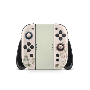 May include: A cream-colored gaming console with black handles and buttons. The left side features a small pagoda and trees, while the right side has floral designs. The center is a light green color.